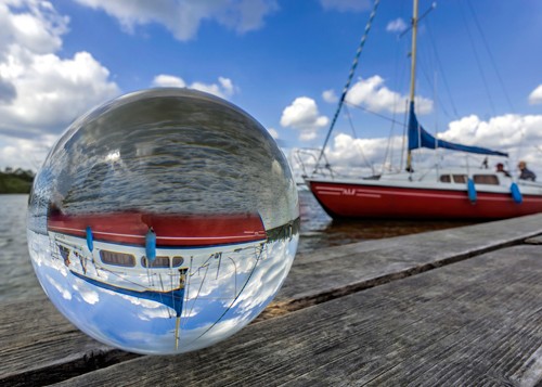 Rolf Fischer, Kugelwelt  Segelboot (Bootssteg, Glaskugel, Kugel,  Segelboot, Himmel, Wolken, Spiegelung, verkehrte Welt, auf dem Kopf stehen, Holzbohlen, Holzsteg, Badezimmer, Arztpraxis, Wohnzimmer, Wunschgröße,  bunt)