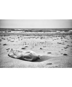 MaciejBledowski, Black and white picture of an empty plastic bottle left on a beach, selective focus, environmental pollution concept picture.